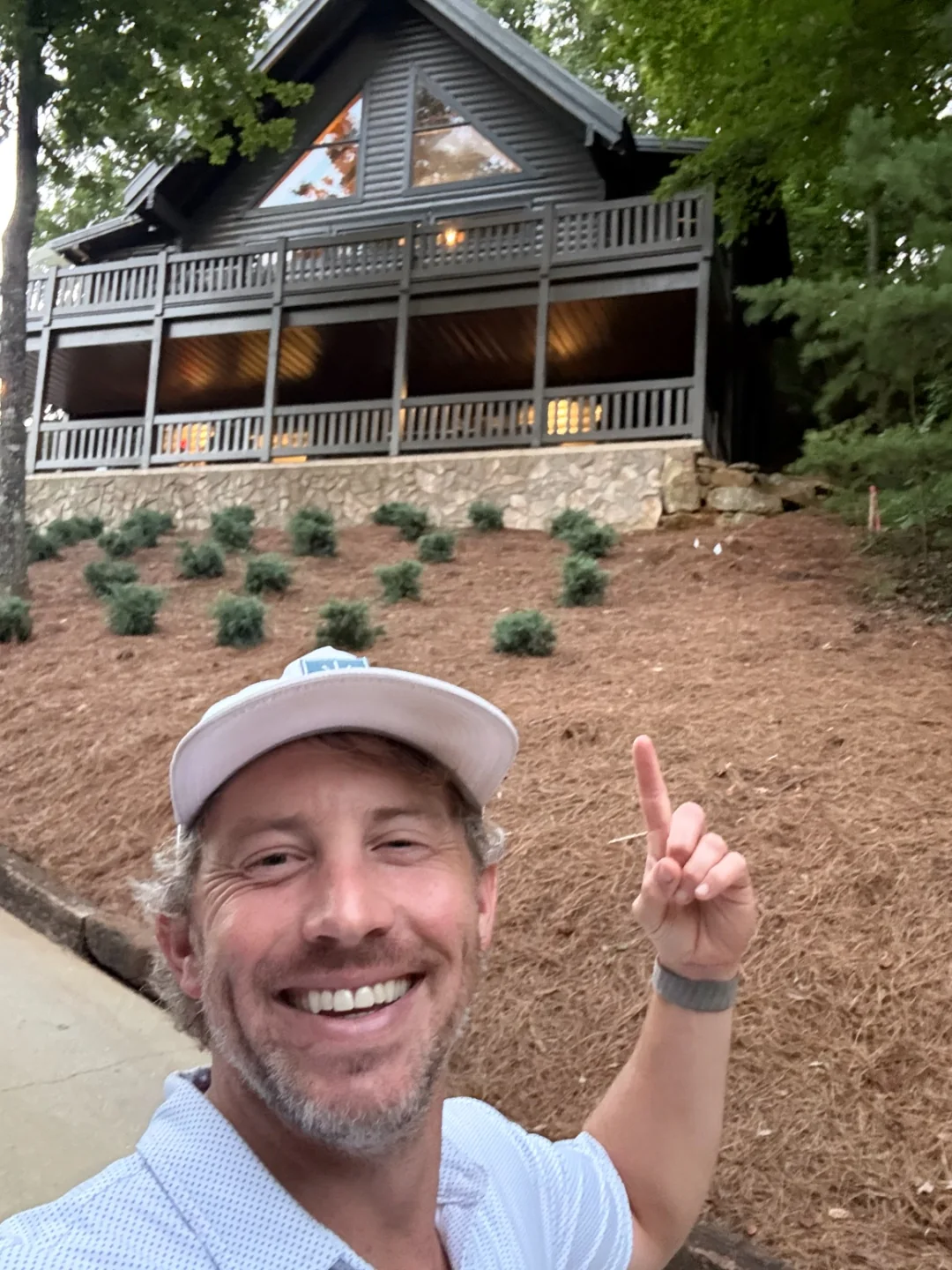 Jeff in a ball cap grinning and pointing up at the lit cabin at dusk on the day they closed on it