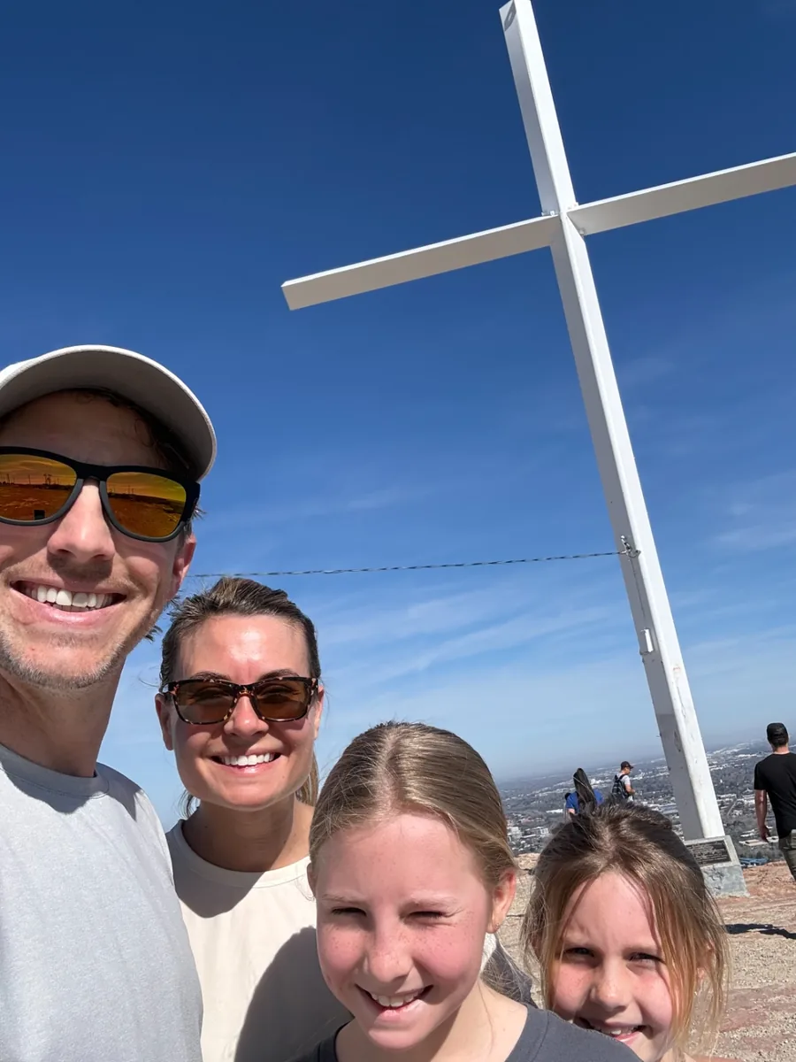Jeff, Chelsea and two of their daughters smiling in a selfie on a sunny hilltop beneath a tall white cross, with the city spread out far below