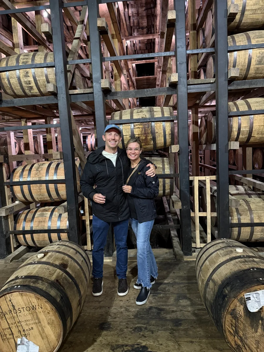 Jeff and Chelsea standing between rows of stacked oak barrels inside a bourbon rickhouse in Kentucky