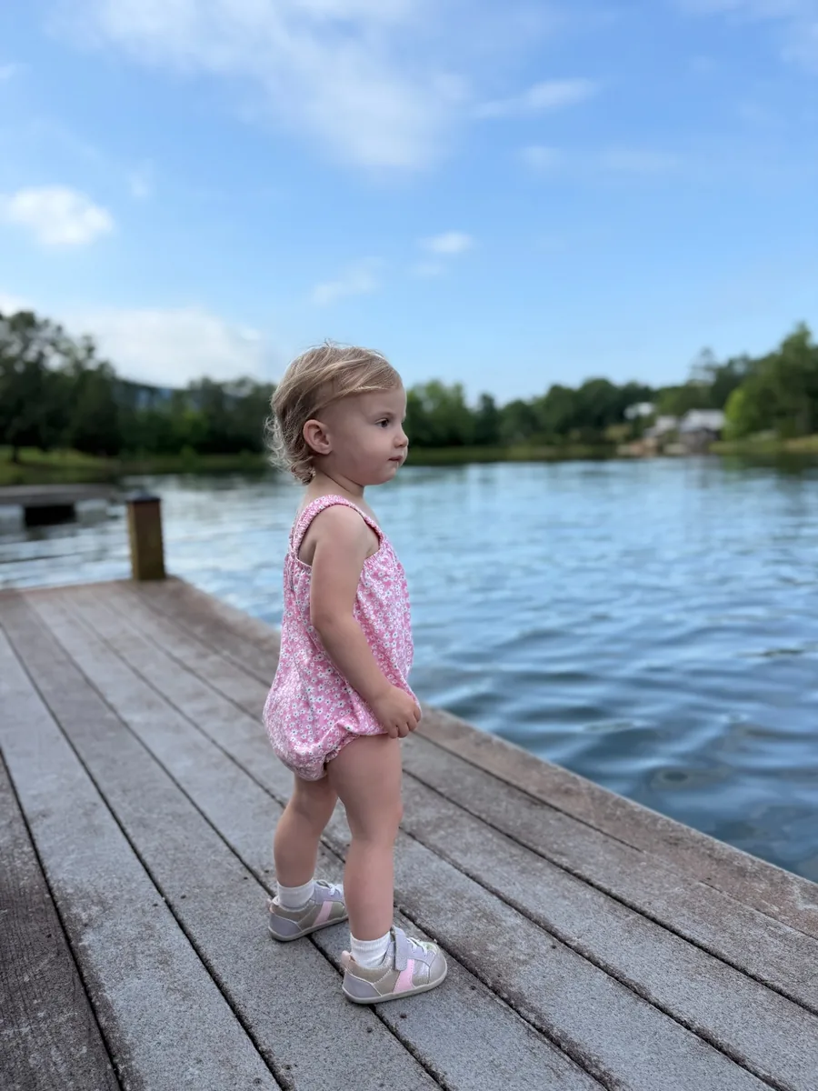 The Mussers' youngest daughter, a toddler in a pink floral romper, standing on a weathered wooden dock looking out over still lake water