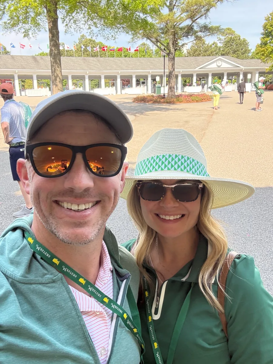 Jeff and Chelsea in green pullovers wearing Masters badges outside the clubhouse at Augusta National, international flags flying behind them