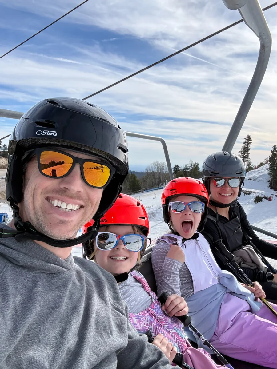 Jeff, Chelsea and two of their daughters in helmets and goggles riding a chairlift up a snowy ski run, one daughter yelling with excitement