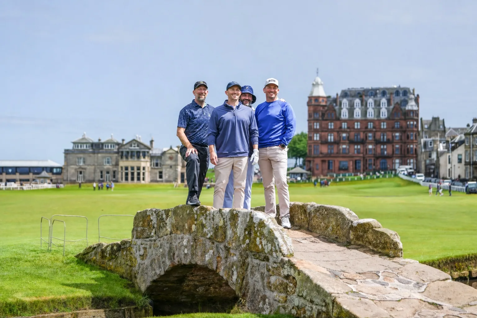 Jeff and three others standing on the stone Swilcan Bridge at the Old Course, St Andrews, with the Royal and Ancient clubhouse and the Hamilton Grand behind them